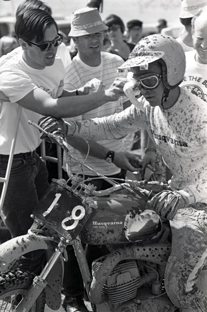 A mud-covered vintage motorcycle racer surrounded by friends and crew.